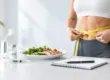 Woman measuring her waist beside a healthy meal and HCG vial during a medically supervised weight loss program.